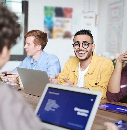 Quatro pessoas estão sentadas em uma mesa com laptops e notebooks, envolvidas em uma discussão. Uma pessoa de jaqueta amarela está falando e gesticulando, enquanto as outras ouvem ou fazem anotações. O cenário parece ser um escritório ou uma sala de reuniões.